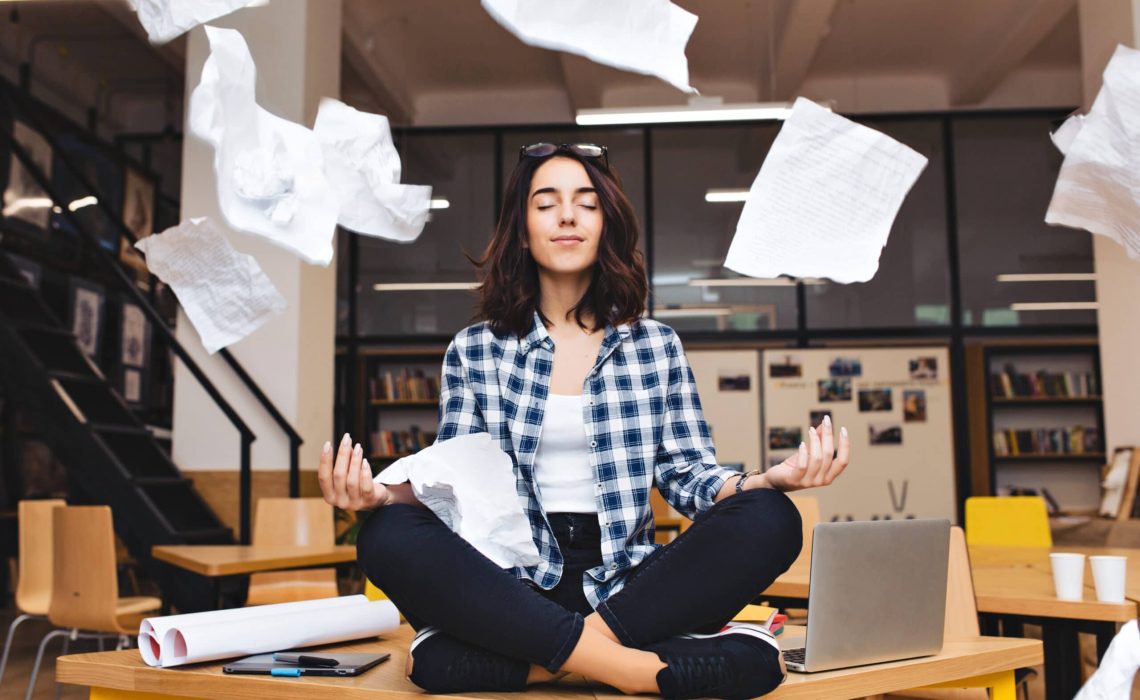 Young pretty joyful brunette woman meditating on table surround work stuff and flying papers. Cheerful mood, taking a break, working, studying, relaxation, true emotions