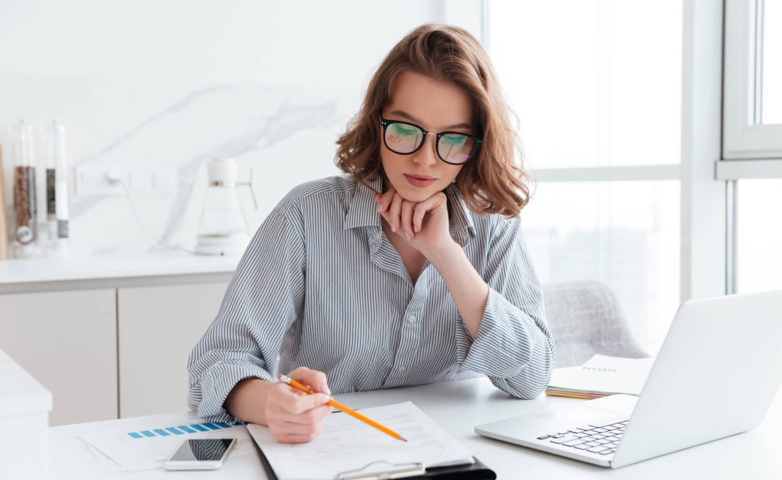 Young concentrated businesswoman in glasses and striped shirt wo Young concentrated businesswoman in glasses and striped shirt working with papers at home