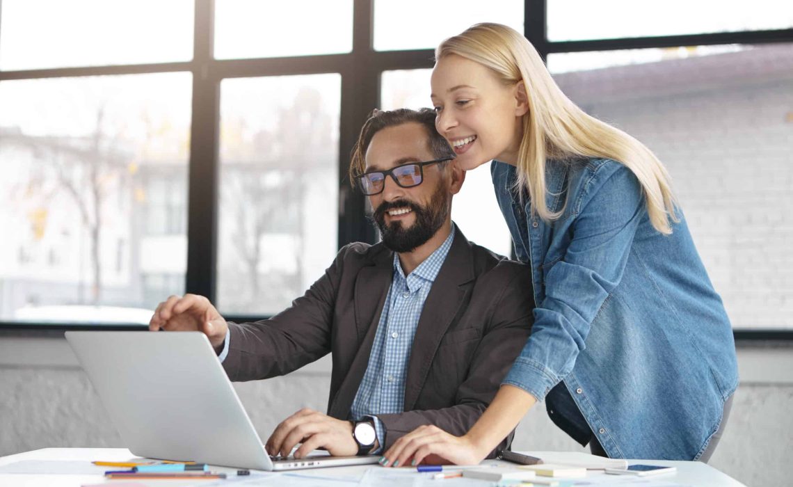 Portrait of successful active coworkers look at laptop computer, sit at work place in office, consult each other what information better to write. Professional investors work on future startup project